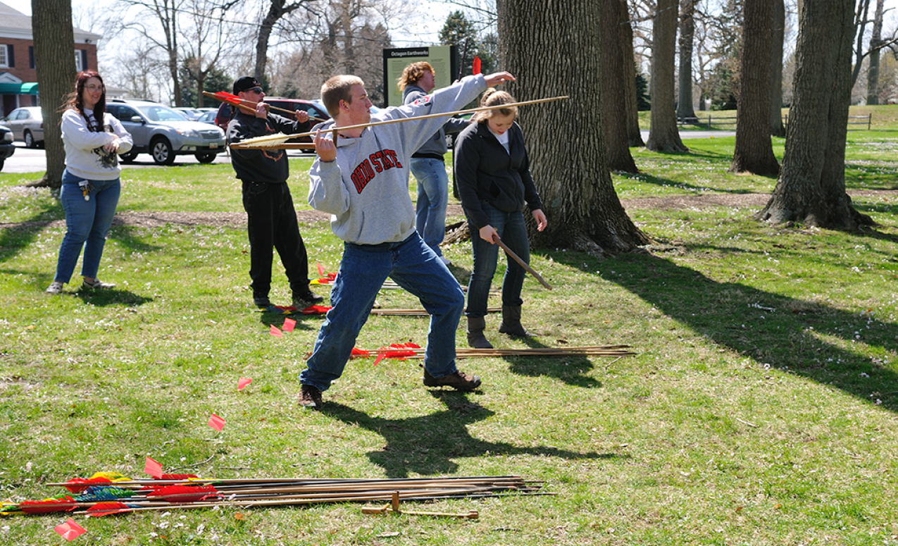Students in mid-motion of throwing an atlatl [an assistive tool for spear throwing] at the Octagon State Memorial, Newark.