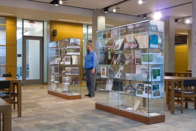 Dr. Richard Shiels standing between two glass exhibition cases of earthworks images and books in the Thompson Library. Image courtesy of The Ohio State University