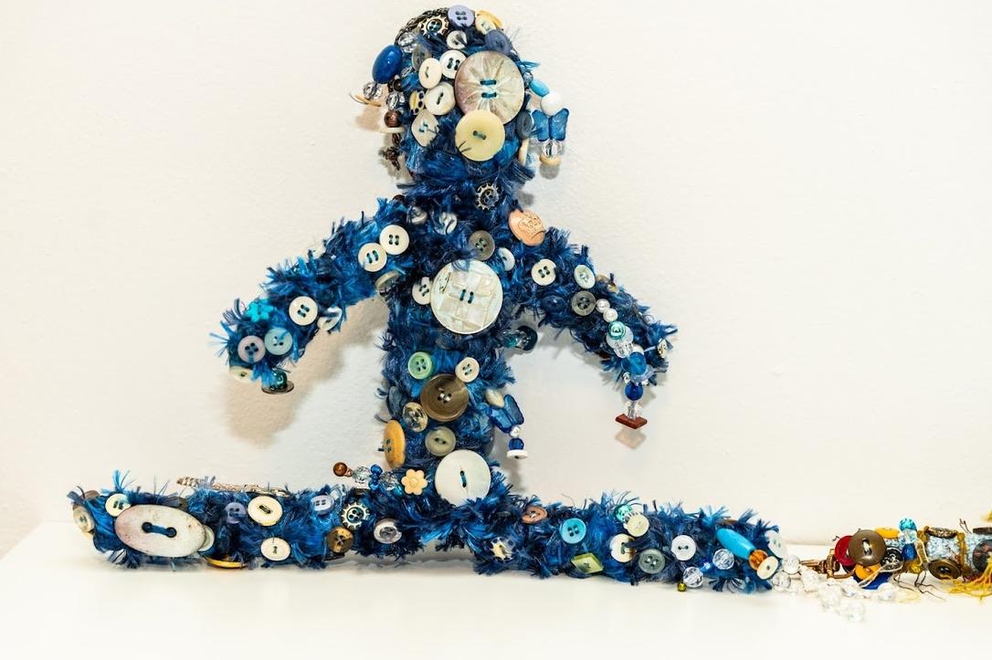 Worry Doll by artist Joy Hoop. A human figure constructed of blue fiber and many colors of buttons doing the splits. Yellow fiber with more buttons transitions to the left of the work.