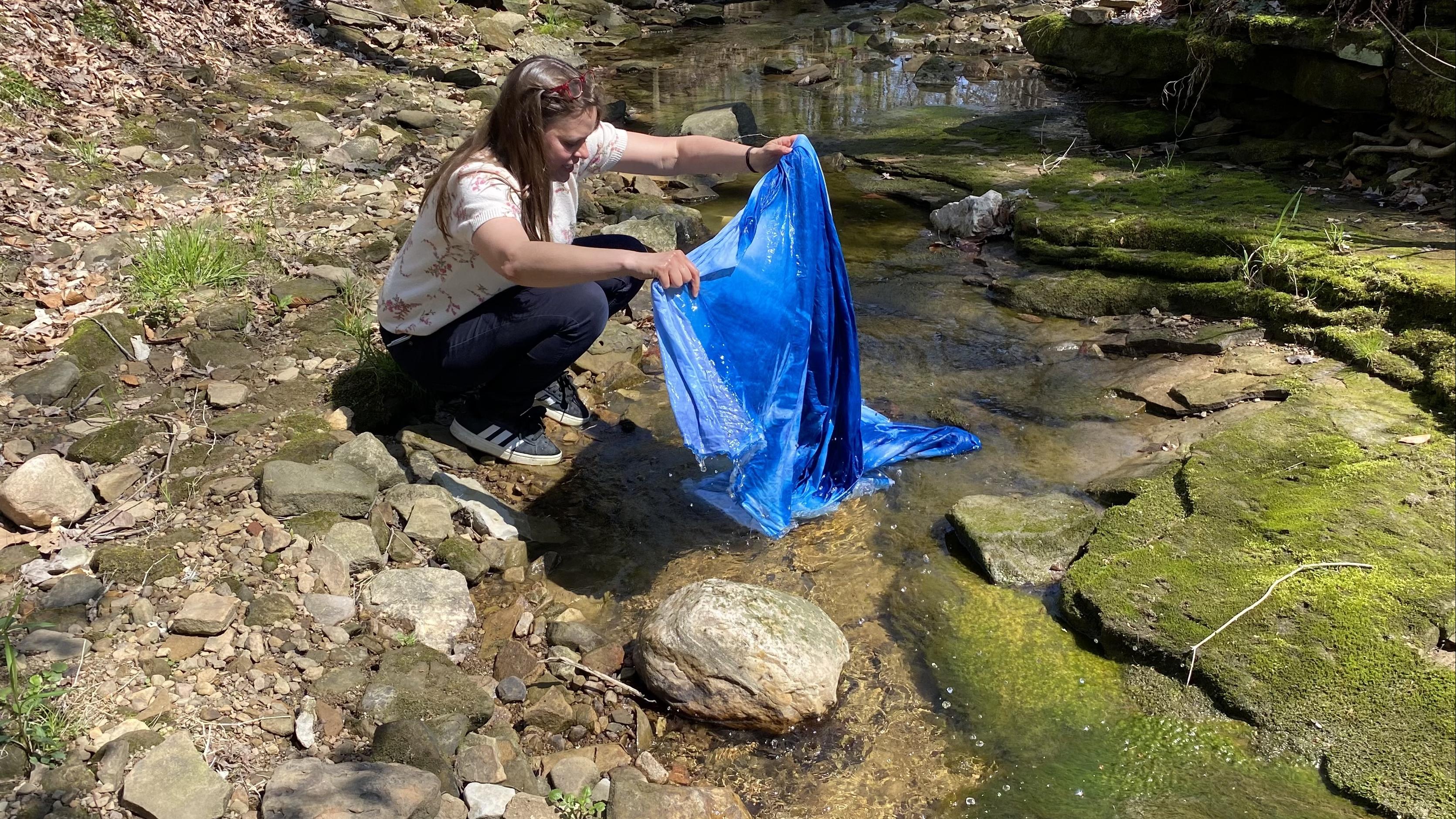 Artist Kate Shannon rinsing a cyanotype printed fabric in a forested creek by a bridge.